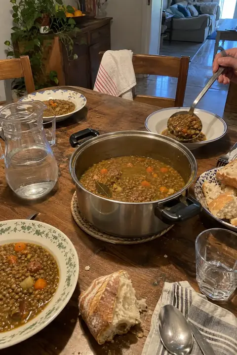 Mesa de casa con una olla de lentejas en el centro y una mano sirviendo con el cucharón